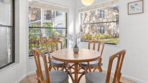 A dining area with a round wooden table and six wicker chairs, large windows with views of greenery and buildings, and a hanging light fixture.