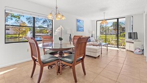 A spacious living area with a dining table, chairs, and a sofa. There are large windows offering a view of the outdoors.