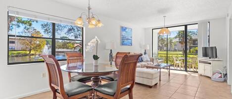 A spacious living area with a dining table, chairs, and a sofa. There are large windows offering a view of the outdoors.