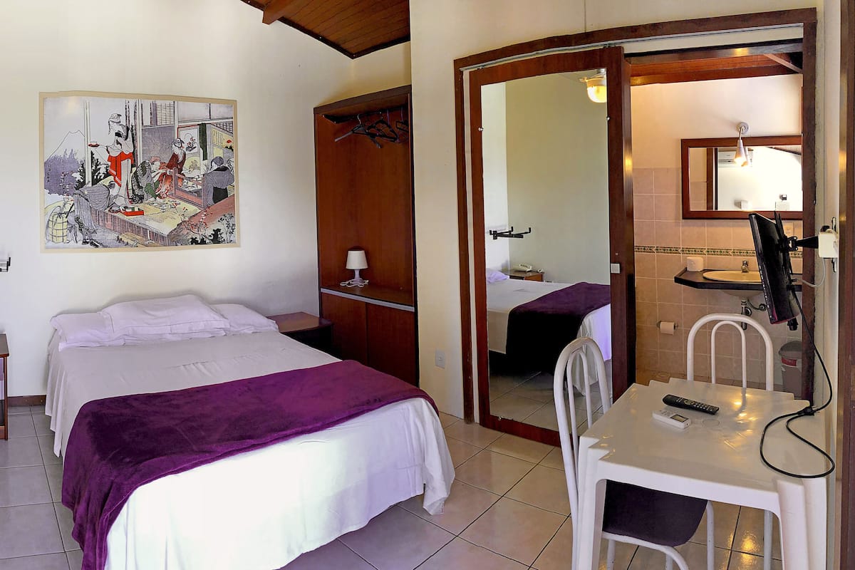A hotel room with a bed, a desk with a chair, a mirror, a painting on the wall, and a bathroom visible through an open door.