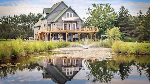 A two-story house with a balcony, a pond in front, and a forested background. - SUMMER SPECIALS  Beautiful lake front, beach and pool table (LaPointe)