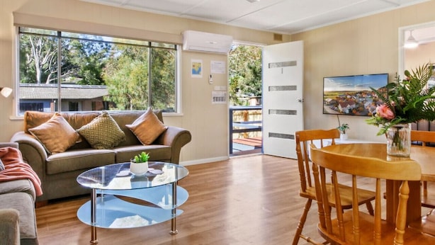 A spacious living area with a sofa, a glass coffee table, a wooden dining table with chairs, a ceiling fan, and a large window offering a view of the outdoors.