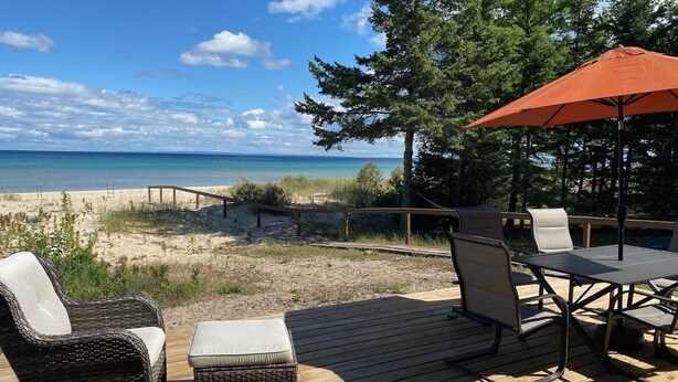 Een houten terras met rieten meubilair, een tafel en een oranje parasol met uitzicht op een strand en een heldere lucht.