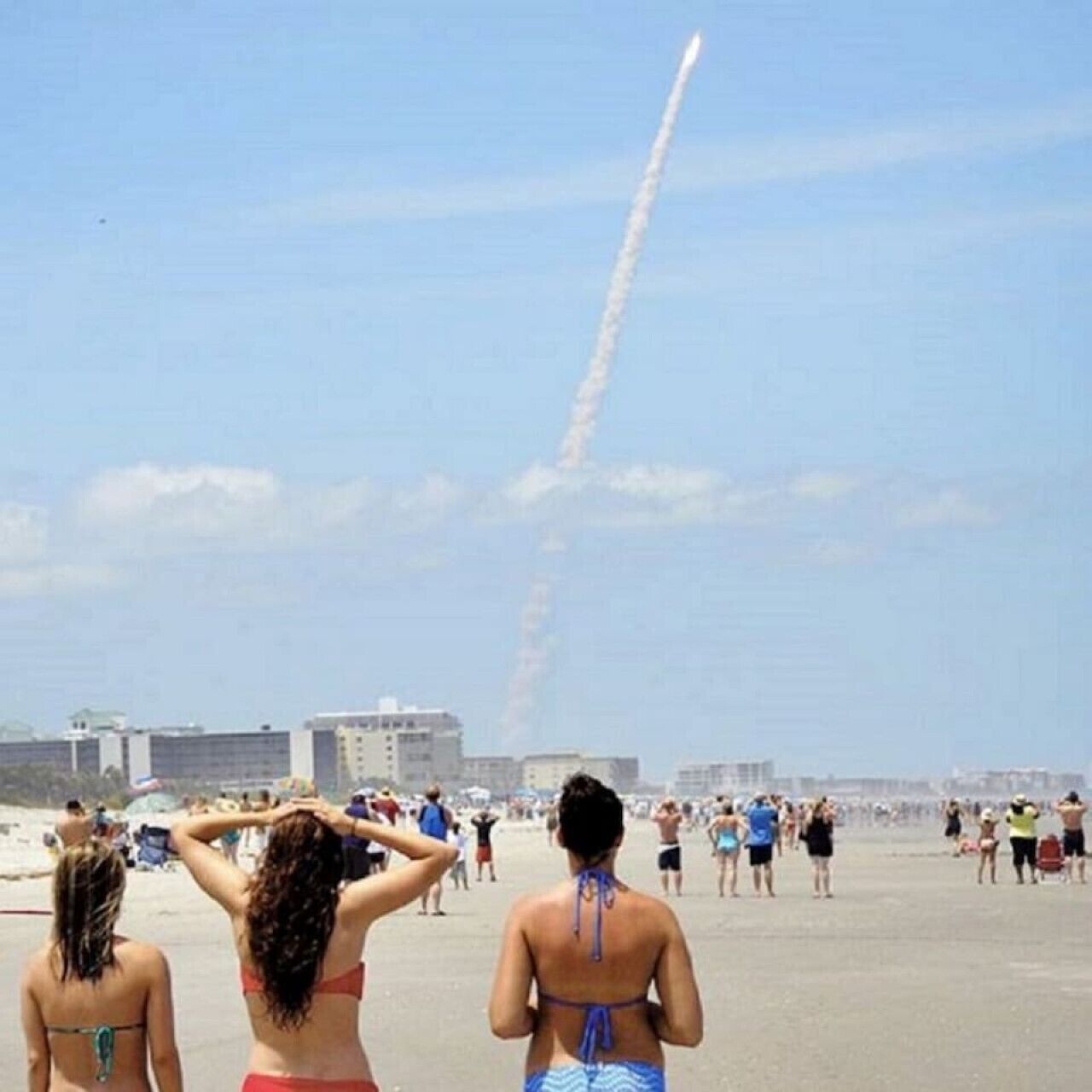 Une plage avec des gens, des bâtiments et une traînée de condensation d’avion visible dans le ciel.