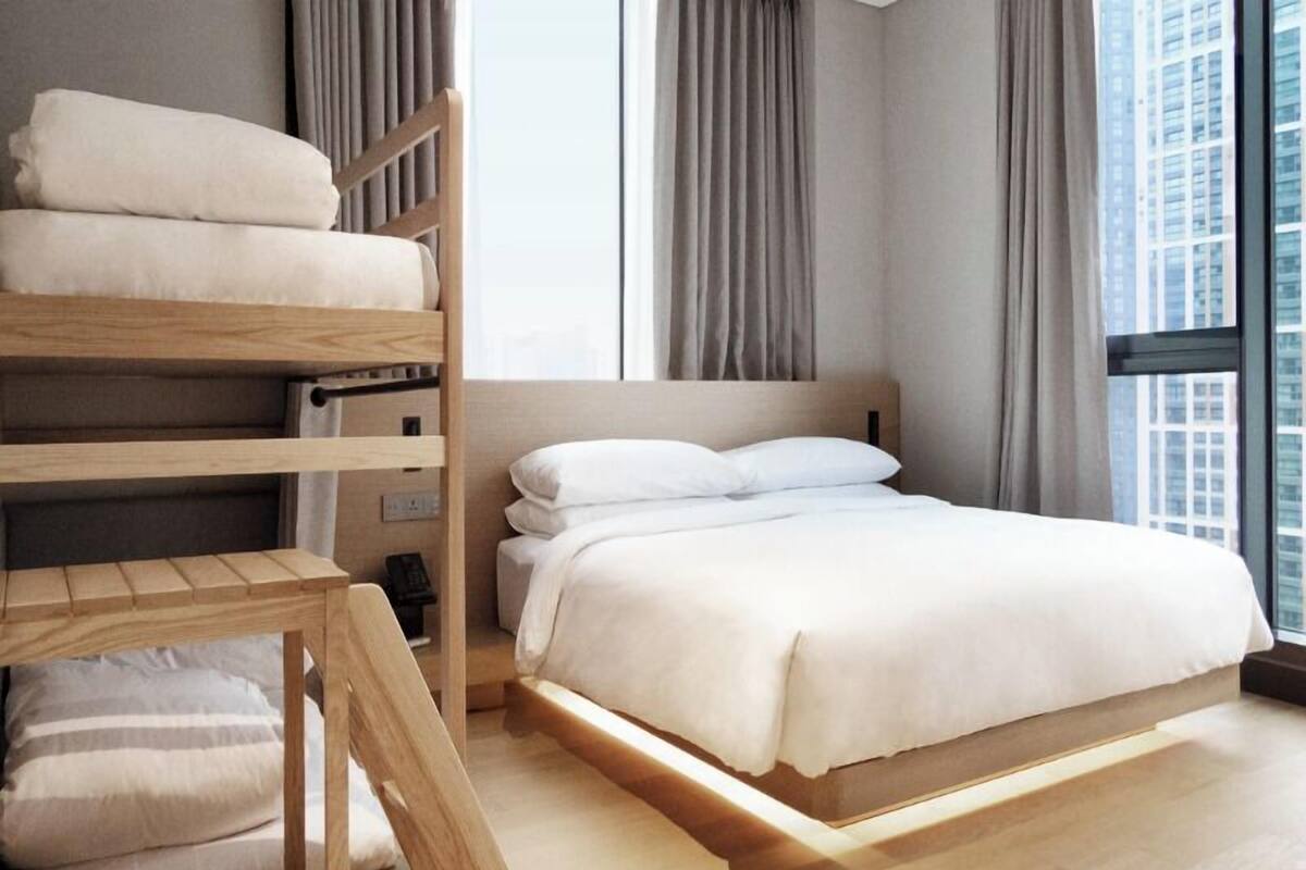 A modern hotel room with a bunk bed, a large bed, and a view of a cityscape through the window.