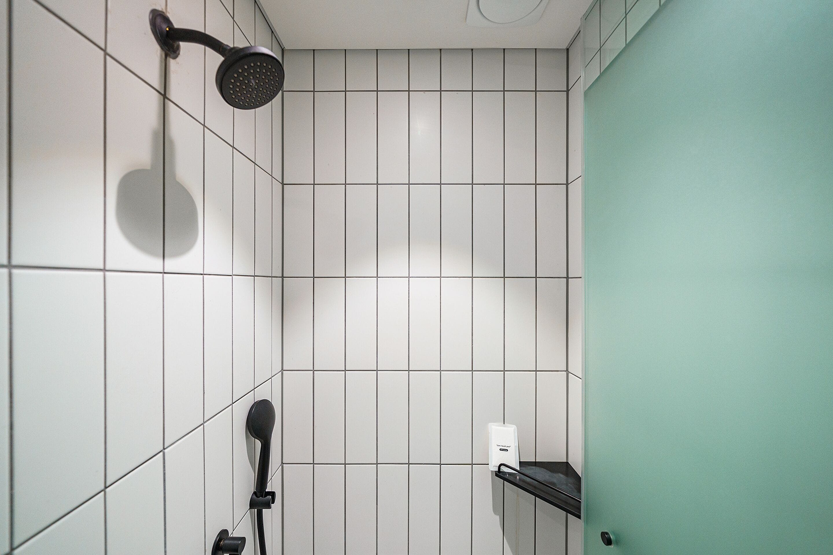 A bathroom with white tiled walls, a black showerhead, and a white soap dispenser on a black shelf.