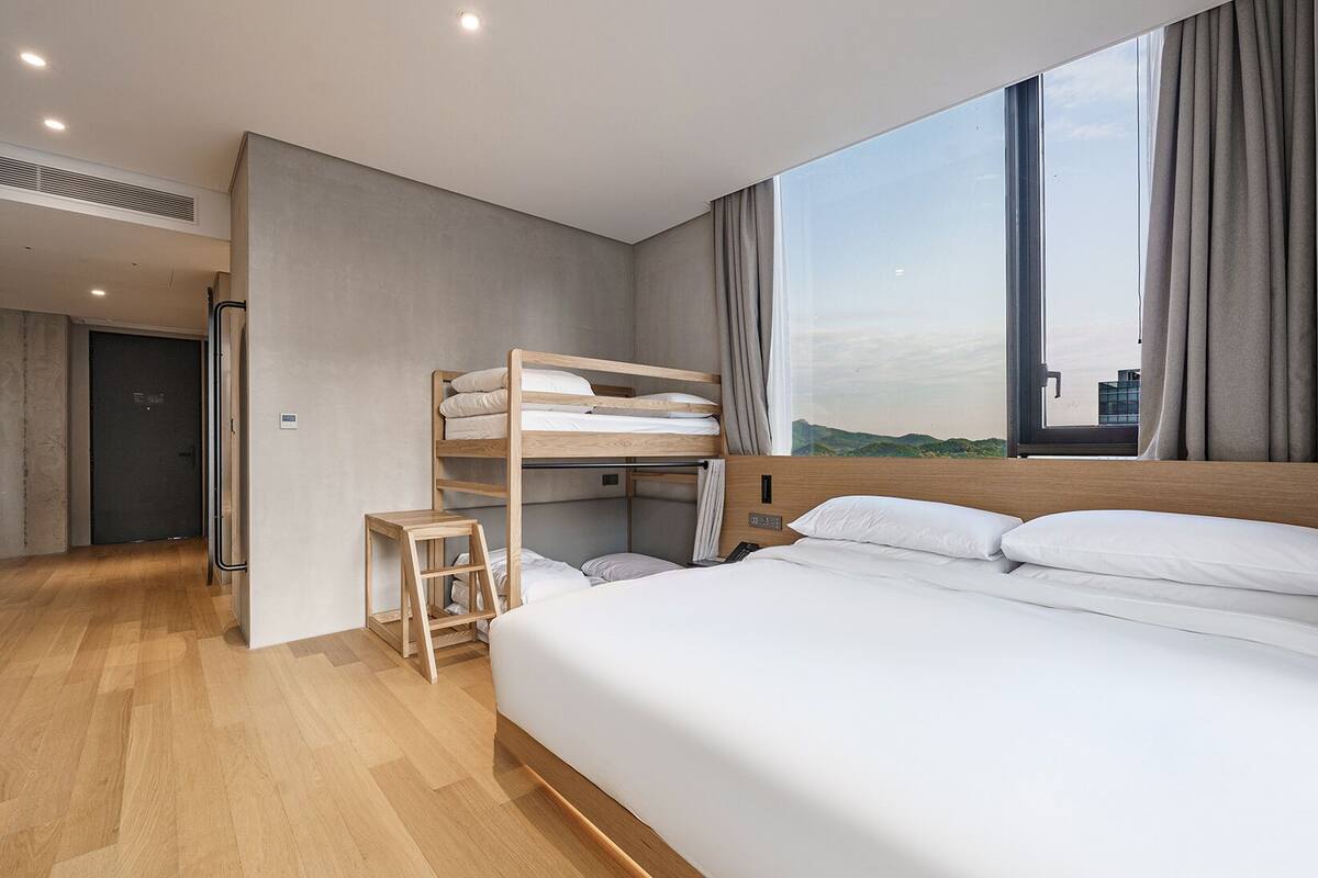 A modern hotel room with a bunk bed, a desk, and a large window offering a city view.