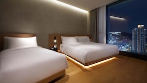 Habitación de hotel con dos camas, una mesita de noche, una lámpara y un ventanal con vistas a la ciudad.