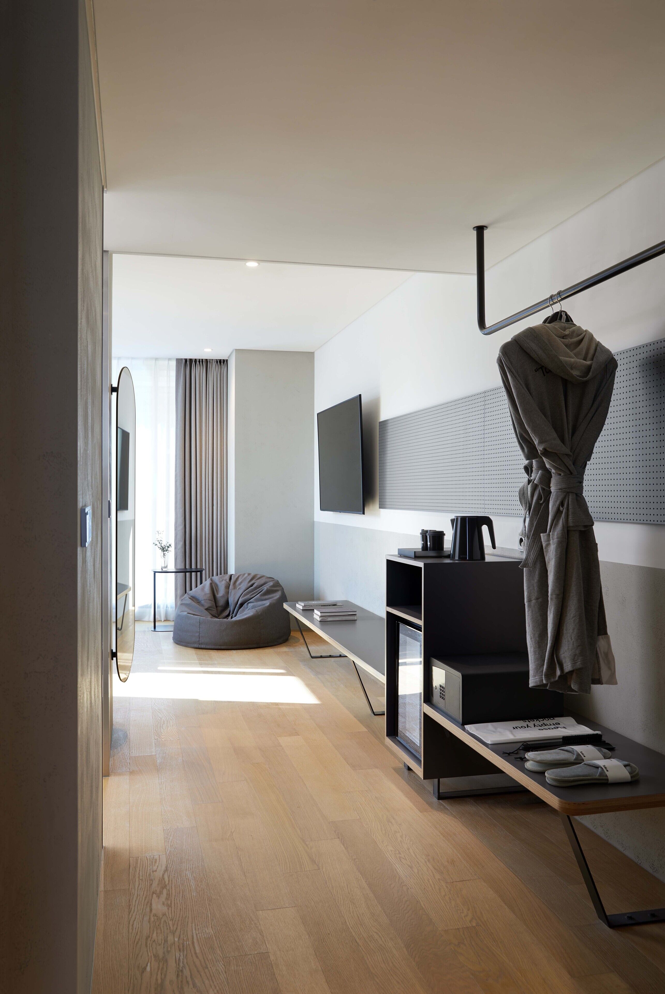 A modern hotel room with a wooden floor, a grey bean bag chair, a flat-screen TV mounted on the wall, a coffee machine, and a robe hanging on a rack.