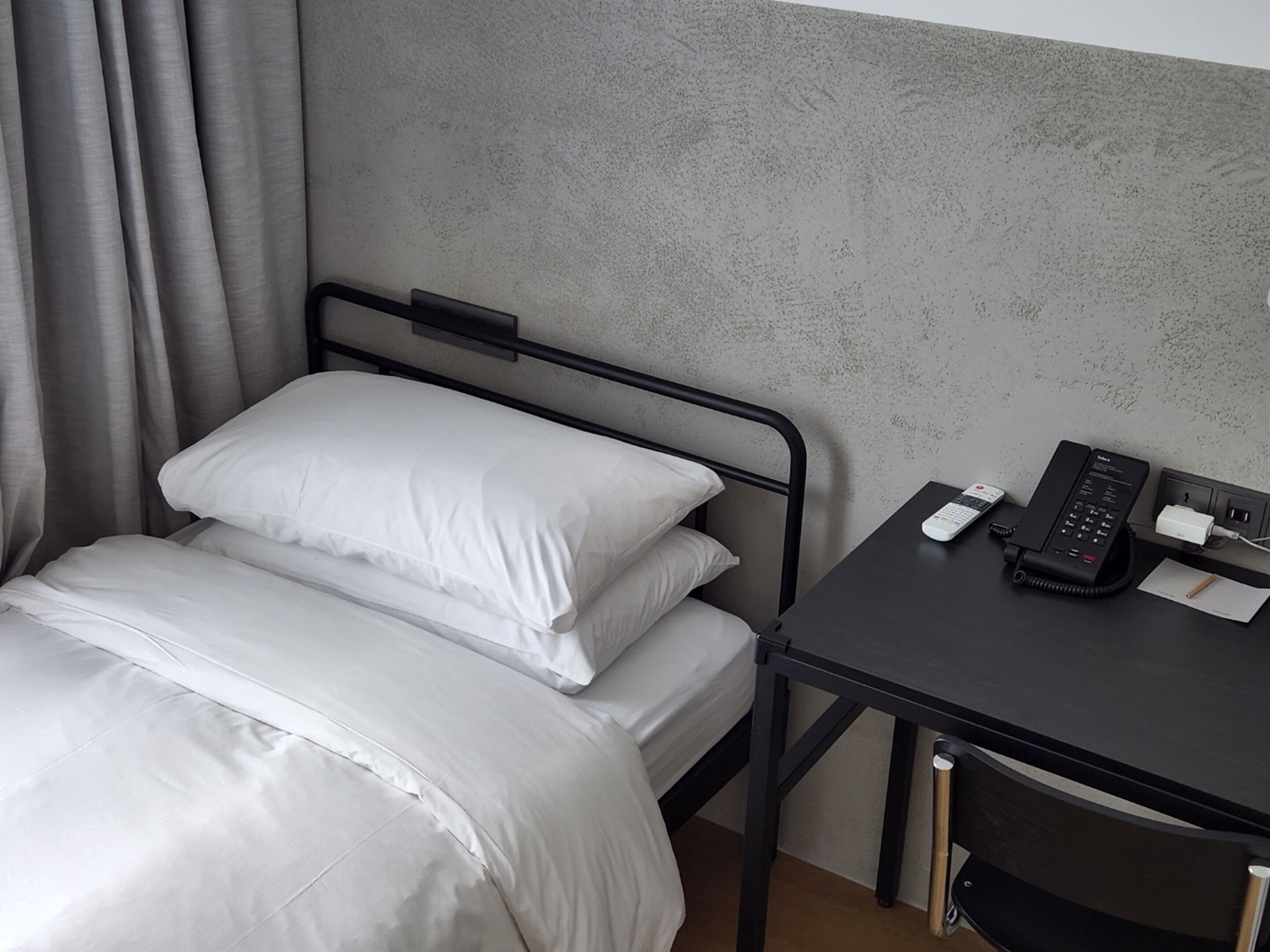 A single bed with white bedding, a black metal headboard, and a desk with a telephone and a power outlet.