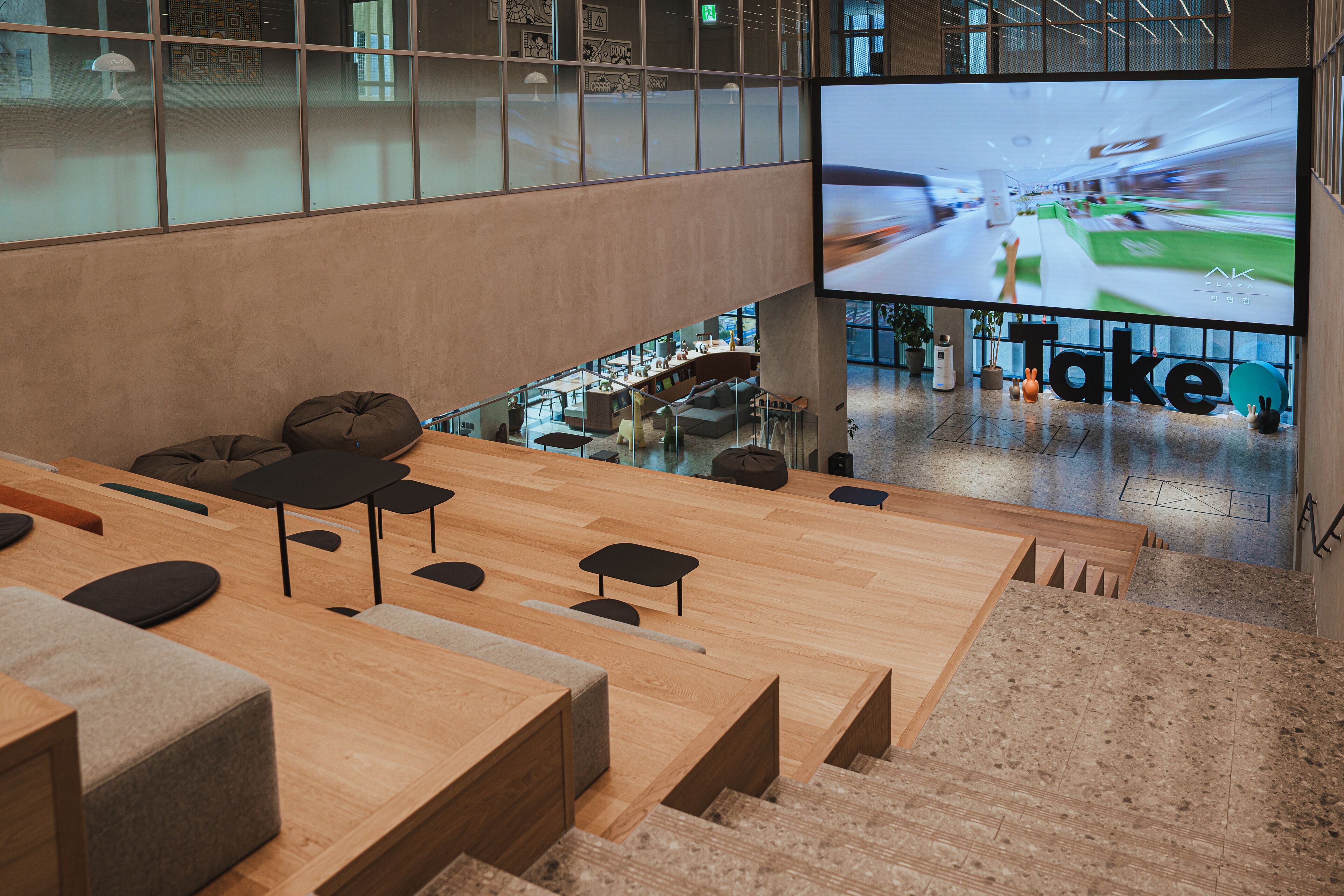 A modern interior with a large screen displaying a sports event, wooden seating areas, and a staircase leading to an upper level.