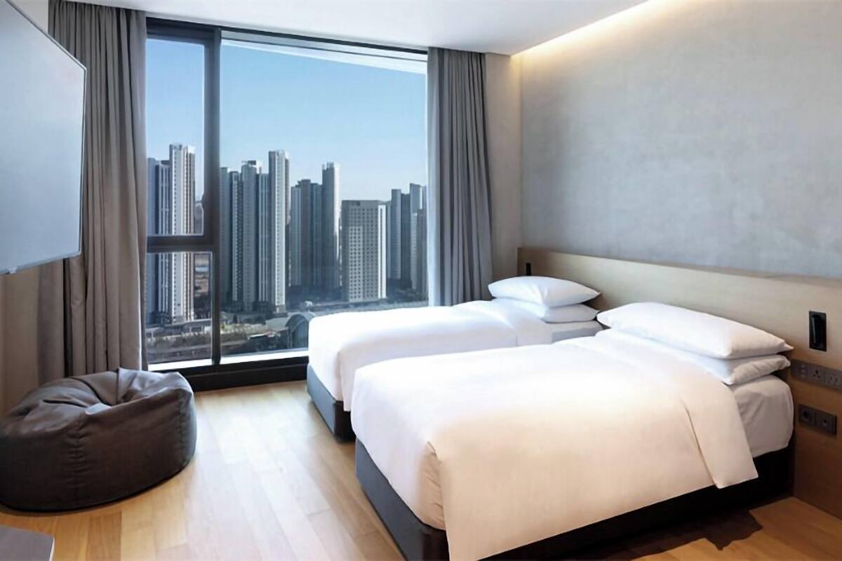 A modern hotel room with a large window, two beds, a bean bag, and a flat-screen TV.