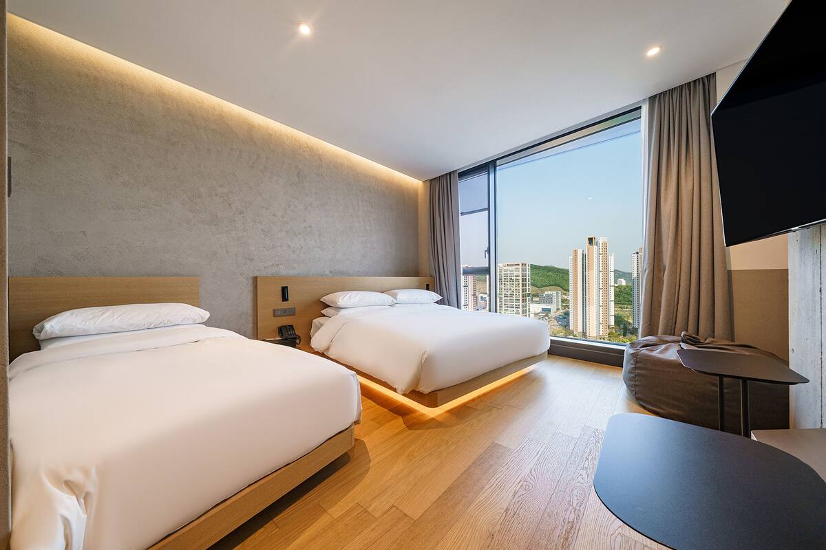A hotel room with two beds, a large window with a city view, a small table, and a flat-screen TV.