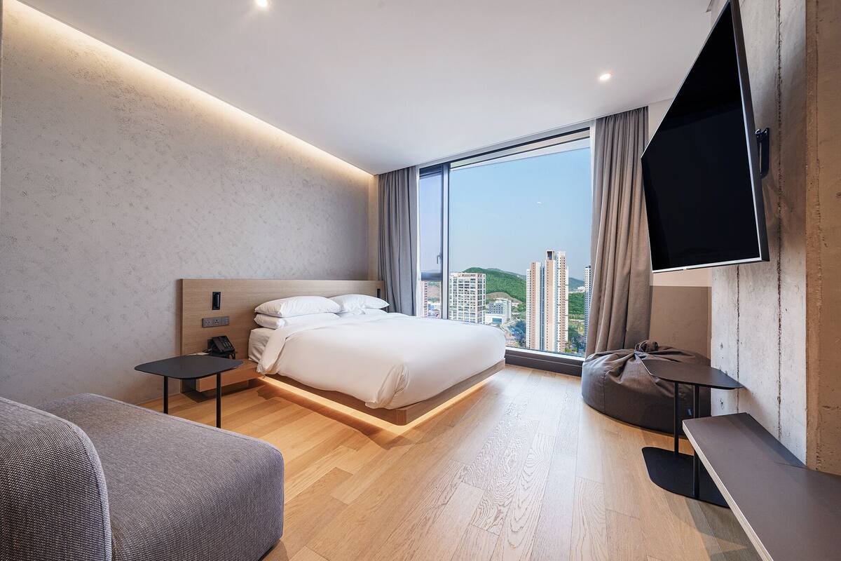 A modern hotel room with a large bed, a desk, a chair, a TV, and a city view.