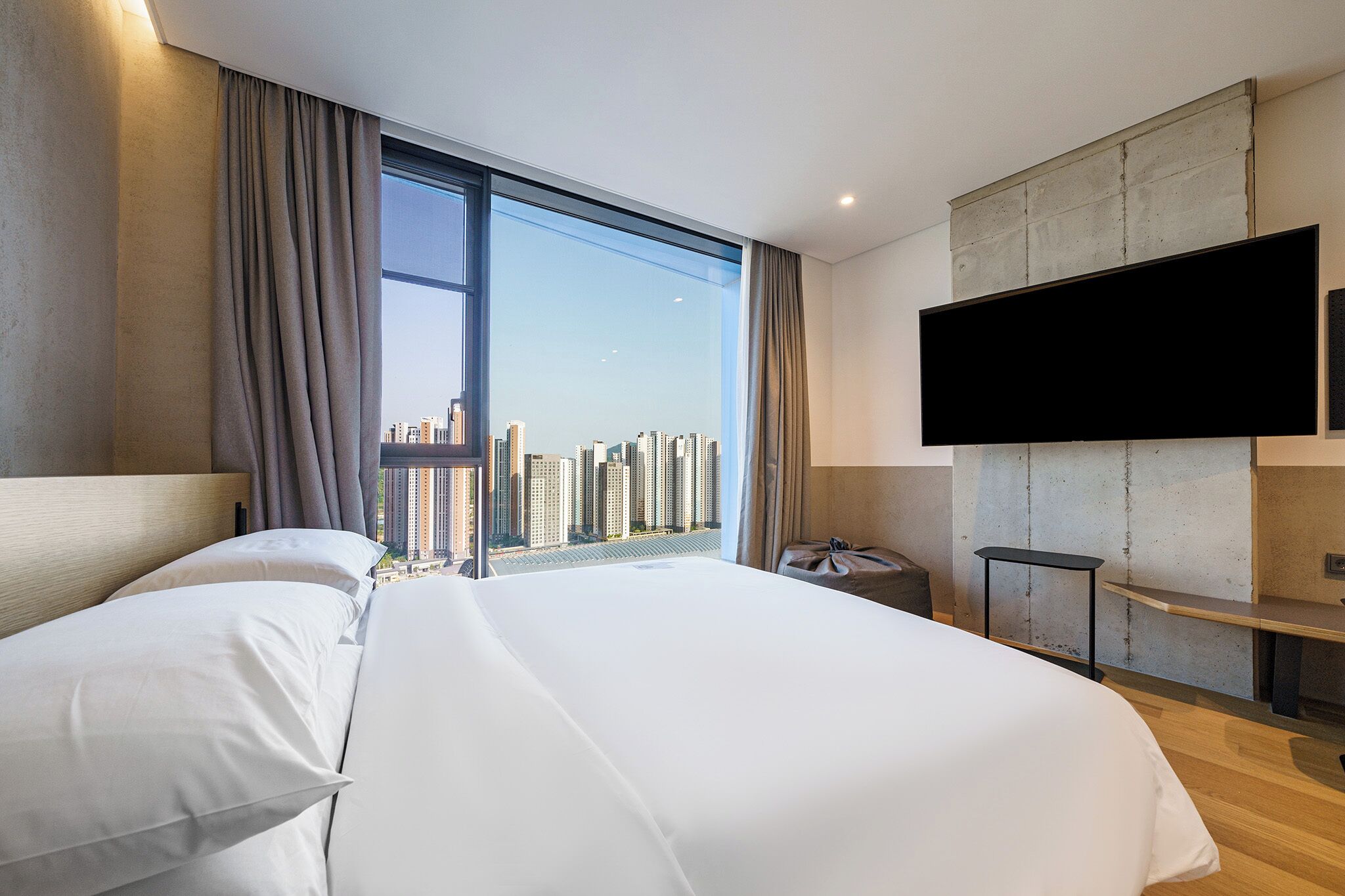 A modern hotel room with a large bed, a flat-screen TV mounted on the wall, and a city view through large windows.