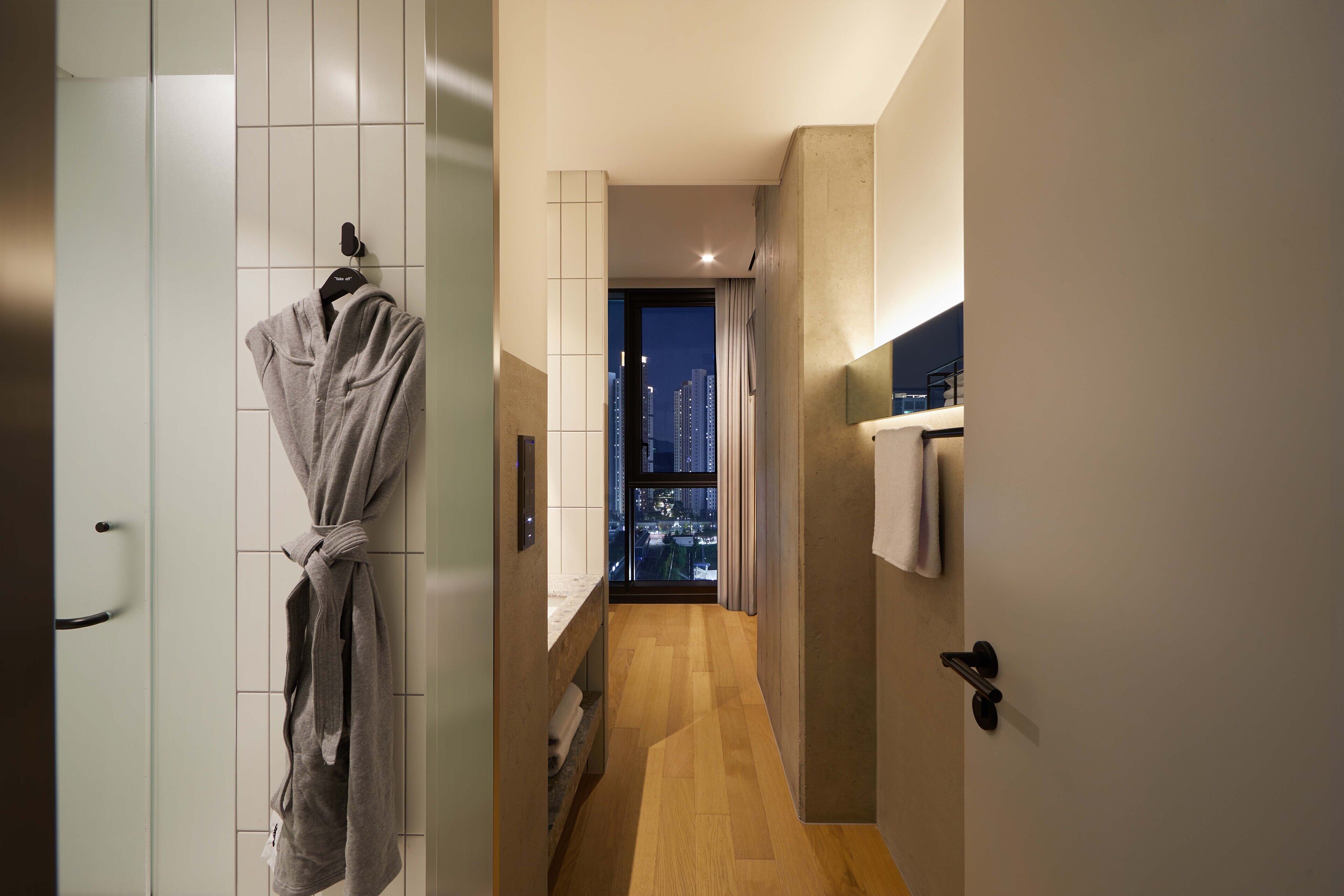 A narrow hallway with a wooden floor, a robe hanging on a hook, and a view of city buildings through a window.