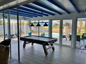 A pool table in a room with large windows, a ceiling fan, and a view of a backyard with a pool. - LILY'S BEACH HOUSE - POOL HOUSE WITH DIRECT RIVER ACCESS (Cape Coral)