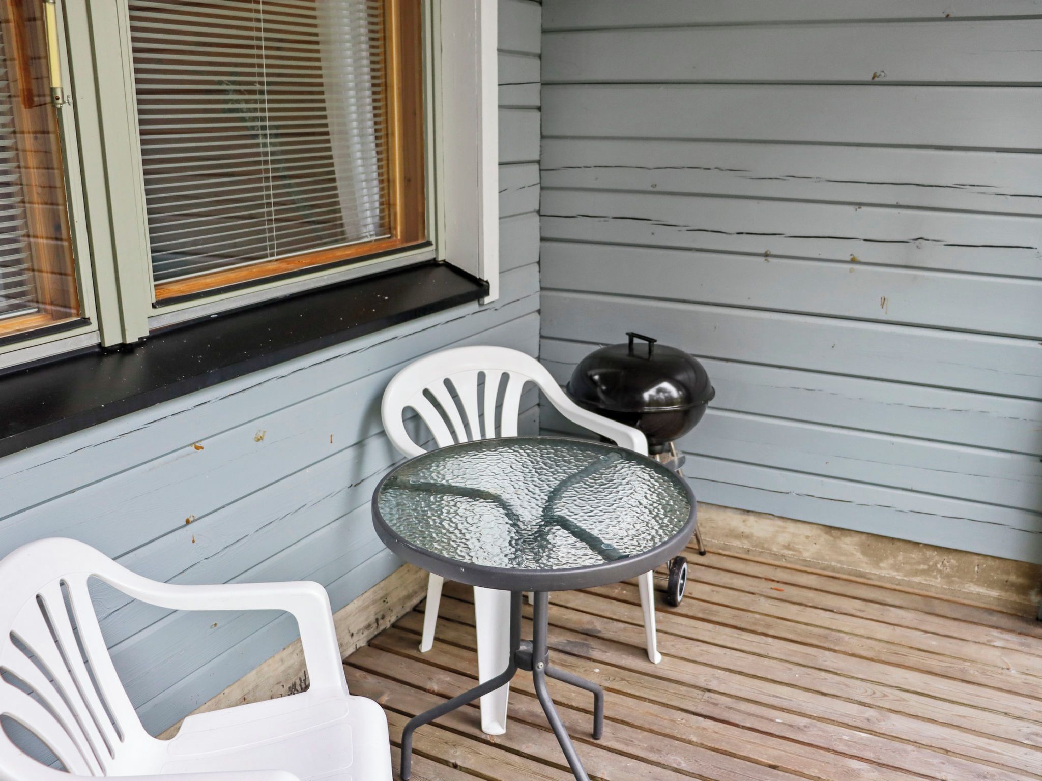 Une véranda avec deux chaises en plastique blanches, une table en verre et un barbecue.