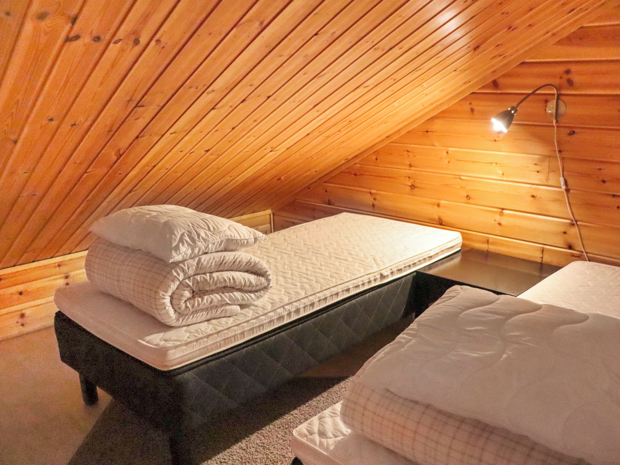 Une petite pièce agréable, avec un plafond en pente, un lit simple avec un matelas et des oreillers, et une lampe de chevet.