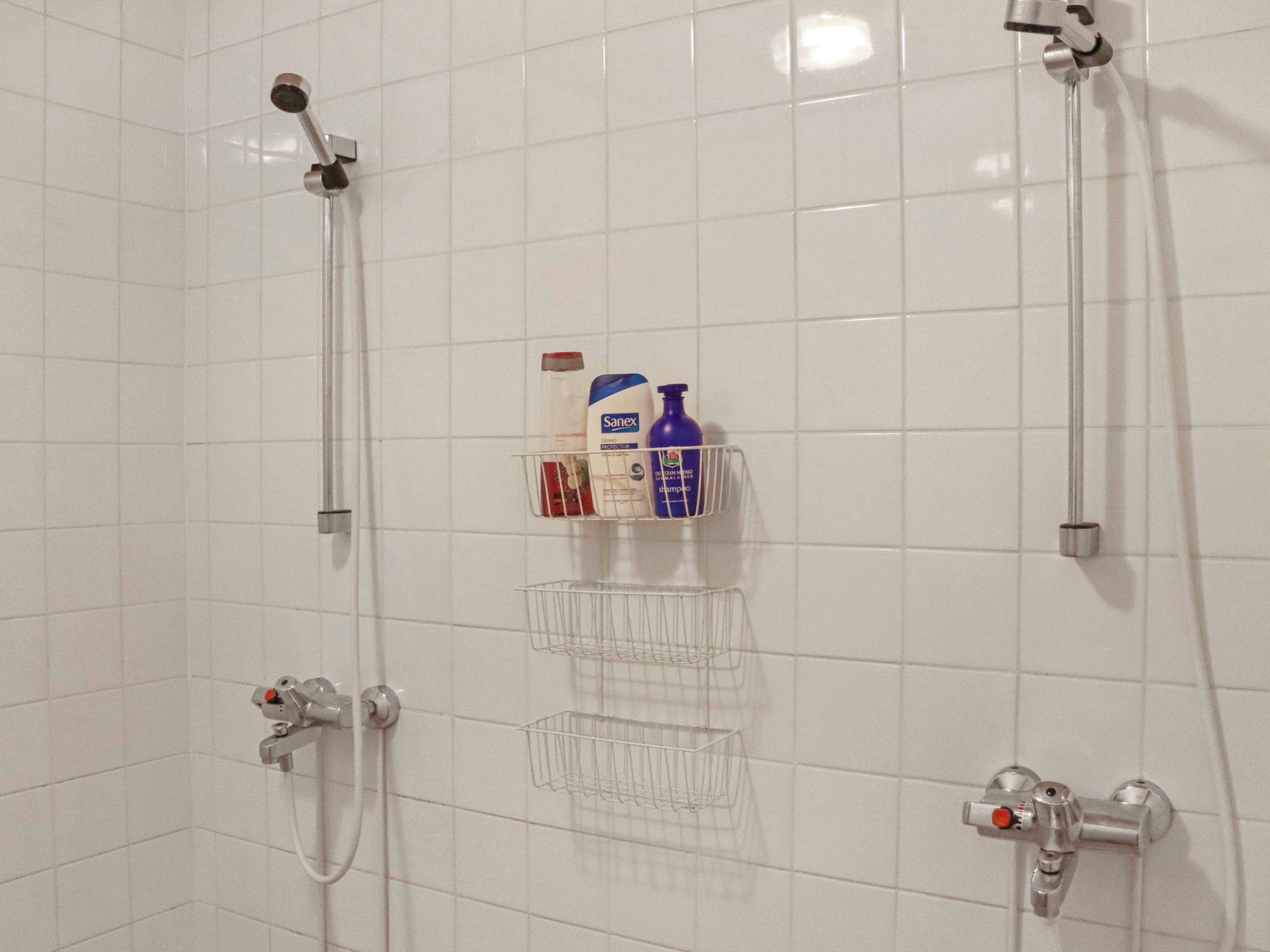 A bathroom with white tiled walls, two showerheads, a white sink with a faucet, and a metal shelf holding toiletries.