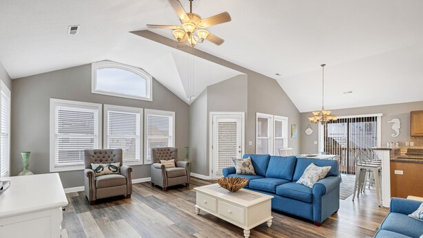A spacious living room with a high ceiling, a blue sofa, two armchairs, a coffee table, and a dining area with a kitchen counter.