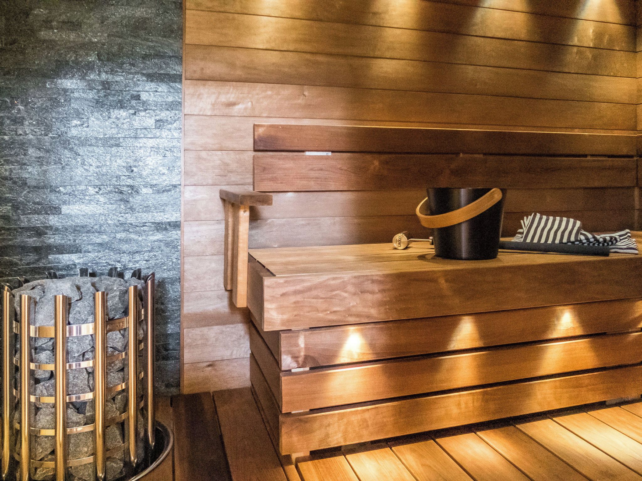 A wooden bench with a black bucket and striped towel, a stone wall, and a metal rack with towels.