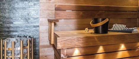 A wooden bench with a black bucket and striped towel, a stone wall, and a metal rack with towels.
