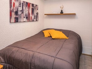 A single bed with a brown duvet and a yellow pillow, a cityscape artwork on the wall, and a shelf with a vase and the word 'Smile'.