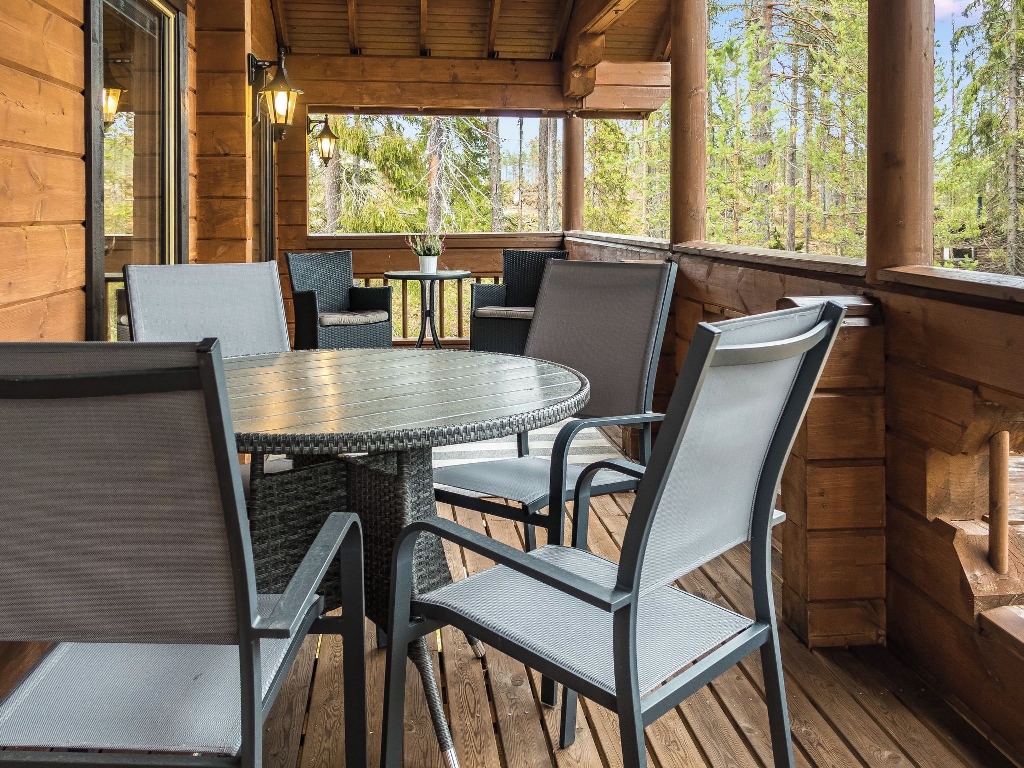 Eine Holzterrasse mit einem runden Tisch und Stühlen, umgeben von Holzwänden und Blick auf Bäume.