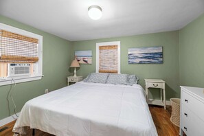 Un dormitorio con cama, mesitas de noche, lámpara, ventana con persianas y obras de arte en las paredes.