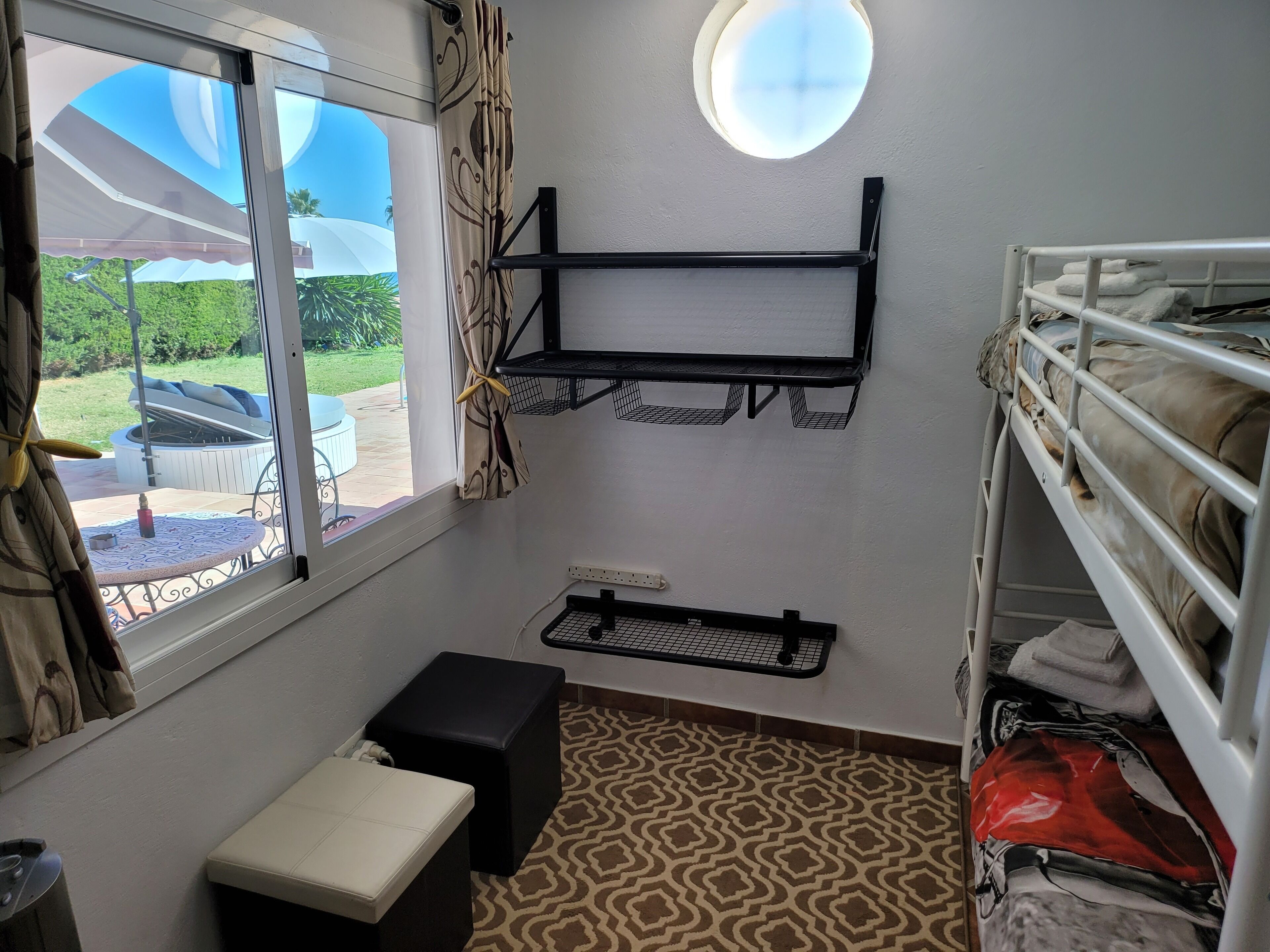 A room with a window, a small table, a bench, a bunk bed, and a wall-mounted shelf.