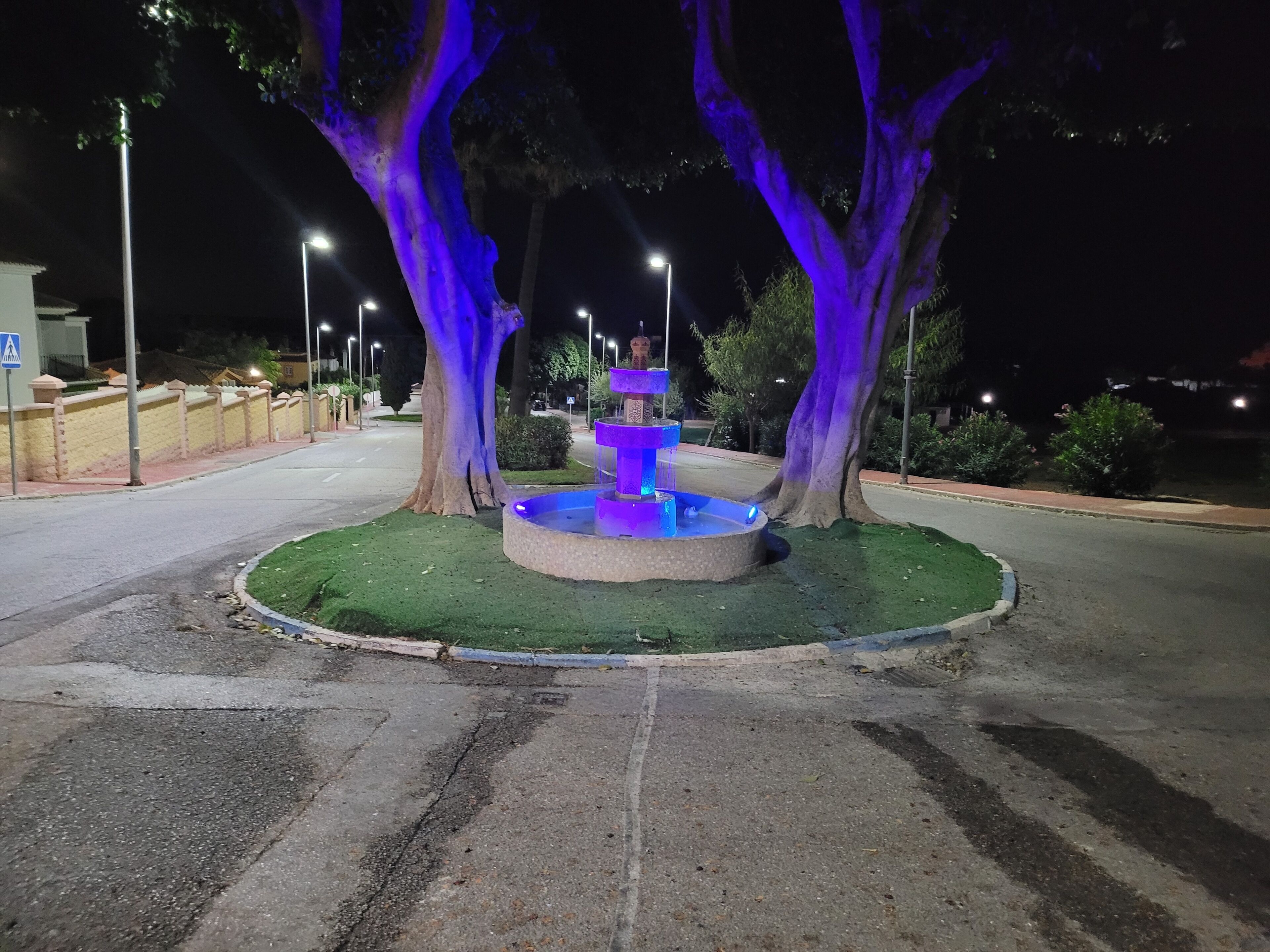 Una fuente bien iluminada con base circular y tres niveles, rodeada de césped y árboles con iluminación azul.
