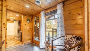 L’intérieur d’un chalet en bois, avec un plancher à carreaux, une chaise en bois, une petite table et une fenêtre avec des rideaux.