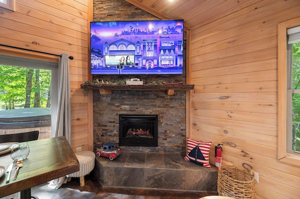 A cozy cabin interior with a stone fireplace, a television mounted above it, and a dining area with a table and chairs.