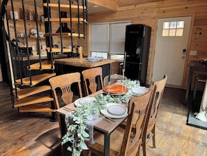 A wooden interior with a dining table set for four, surrounded by wooden chairs. A spiral staircase with black metal railings is visible in the background.