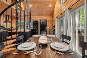A wooden cabin interior with a dining table set for four, a kitchen area, and a spiral staircase. - MoonLight Treehouse | Hot Tub · Lakefront Retreat (Belgrade)