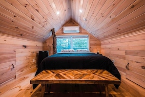 A cozy wooden cabin room with a bed, a table, and a window providing a view of trees.