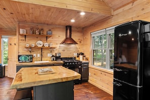 A wooden cabin interior with a kitchen featuring a black refrigerator, a stove, and wooden shelves with decorative items.