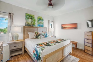 A bedroom with a bed, nightstands, a desk, a TV, artwork, and a view of the outdoors.