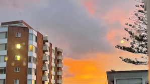 A residential street with a row of parked cars, a tall apartment building, and a sunset in the background. - Spacious Manly Studio (Fairlight)