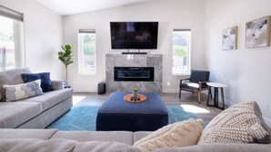 Ein modernes Wohnzimmer mit einem flachbildfernseher über einem Kamin, einem grauen Sofa, einem blauen Hocker und einem kleinen Beistelltisch.