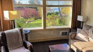 Hotelkamer met een groot raam met uitzicht op een tuin en bomen. Er staat een bruine leren fauteuil, een beige bank en een kleine ronde tafel met een lamp.