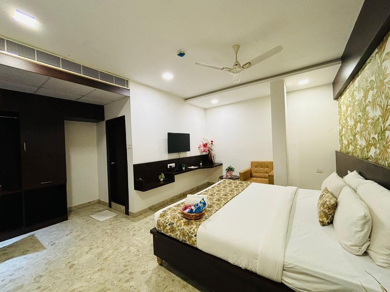 A hotel room with a bed, a TV mounted on the wall, a ceiling fan, and a small table with a vase of flowers.