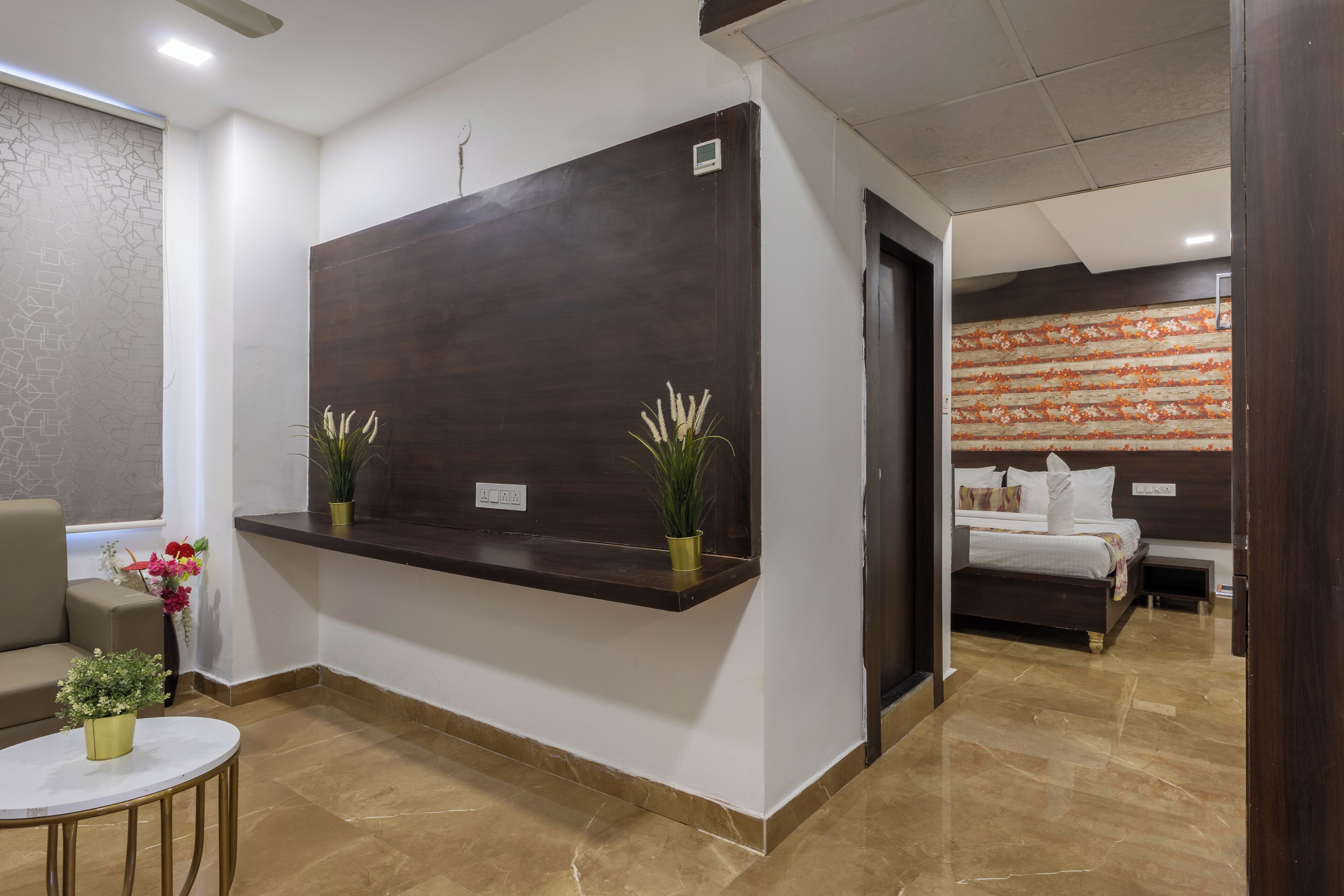 Zona de recepción de hotel con un mostrador de madera oscura, un sofá, una mesita redonda con una planta y una cama al fondo.