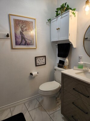 Un bagno con il water, un lavandino con uno specchio, un quadro incorniciato a parete, un mobile con piante e un cartello con un messaggio divertente.