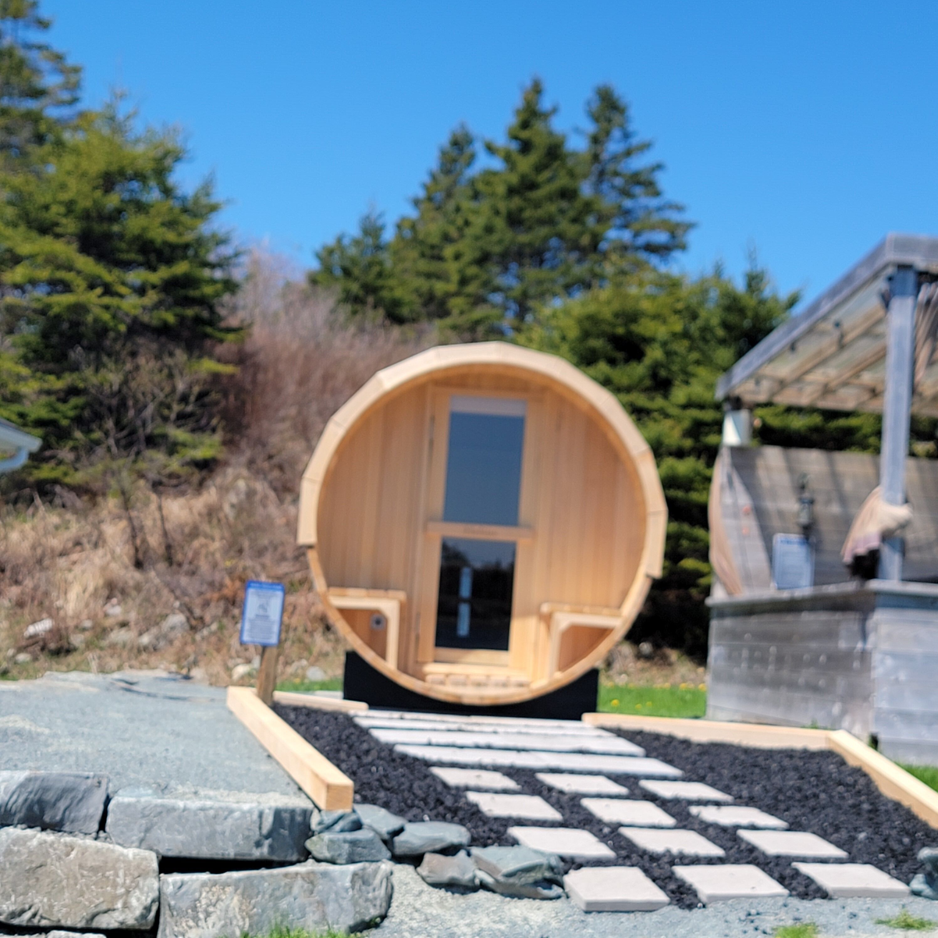 A wooden sauna structure with a door, surrounded by a stone pathway, and a signboard in the foreground.