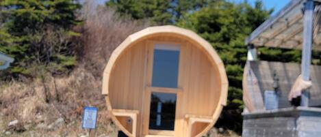 A wooden sauna structure with a door, surrounded by a stone pathway, and a signboard in the foreground.