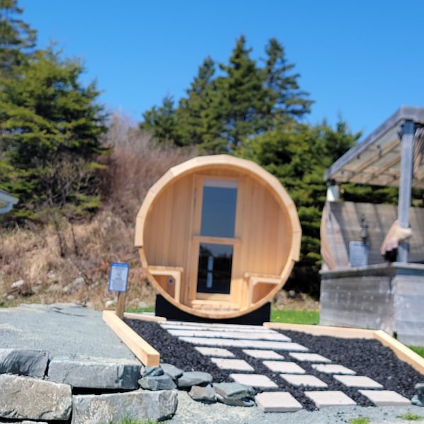 A wooden sauna structure with a door, surrounded by a stone pathway, and a signboard in the foreground.