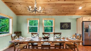 A dining area with a wooden table and benches, large windows with a view of greenery, and a chandelier. - New ModernRustic House in Smokys-4BR/Pools/Hot Tub/Billards/Games (Gatlinburg)