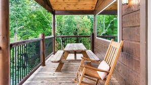 A wooden deck with a picnic table and a rocking chair, surrounded by a wooden railing and a view of a forest. - New ModernRustic House in Smokys-4BR/Pools/Hot Tub/Billards/Games (Gatlinburg)